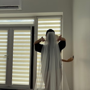 May include: A long, sheer white bridal veil. The veil is draped over the head and shoulders. The person is wearing a black top. The background includes a window with blinds and a white wall.