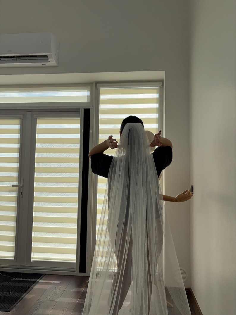 May include: A long, sheer white bridal veil. The veil is draped over the head and shoulders. The person is wearing a black top. The background includes a window with blinds and a white wall.