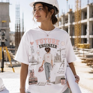 May include: A white t-shirt featuring a graphic print with the text "FUTURE ENGINEER" and illustrations of a person, a bridge, a crane, a laptop, books, and engineering tools. The design includes pink and black accents on a white cotton fabric.