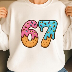 May include: White sweatshirt featuring a colorful donut design with the number 67. The number 6 is pink with sprinkles, and the number 7 is blue with sprinkles. The donuts have a brown base.