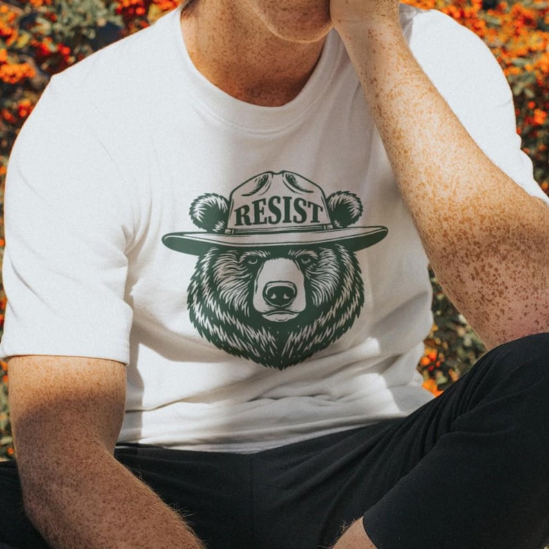 May include: White t-shirt with a green bear wearing a ranger hat that says "RESIST" on it.