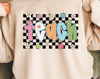 May include: Beige sweatshirt with the word "TEACH" in colorful, patterned letters. The "T" is pink with a leopard print, the "E" is white with a star, the "A" is orange with a checkered pattern, the "C" is blue with stripes, and the "H" is green with stars. The word is set against a black and white checkered background with flowers.