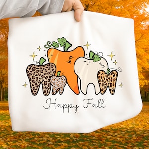 Peut inclure: Sac fourre-tout blanc avec un motif automnal fantaisiste. Il présente des dents et des citrouilles, avec des imprimés léopard sur certaines dents et des citrouilles orange. Le texte "Happy Fall" est écrit en cursive. Des étoiles dorées parsèment le motif.