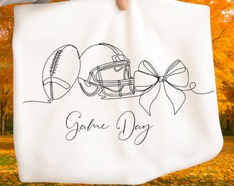 May include: White fabric with a black line drawing of a football, helmet, and bow. The words "Game Day" are written in a cursive font below the drawings. The background is a blurred image of fall foliage.