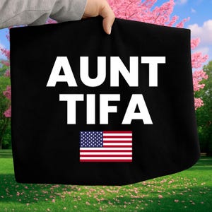 May include: Black tote bag with the text "AUNT TIFA" in large white letters and a small American flag graphic. The bag is held up against a background of green grass and pink flowers.