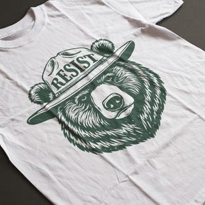 May include: White t-shirt with a green graphic of a bear wearing a ranger hat with the word "RESIST" on it.
