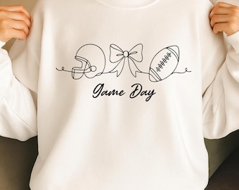 May include: White sweatshirt with a black line drawing of a football helmet, bow, and football. The words "Game Day" are written in a script font below the drawing. The sweatshirt is a casual, comfortable style.