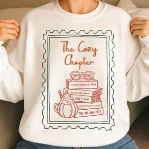 May include: White sweatshirt with a graphic design featuring the text "The Cozy Chapter" in brown. The design includes a stack of books, glasses, and a pumpkin spice latte illustration in shades of brown and orange.