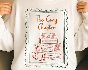Może przedstawiać: Biała bluza z nadrukiem w kolorze brązowo-pomarańczowym. Projekt zawiera napis "The Cozy Chapter", stos książek, okulary i ilustrację pumpkin spice latte.