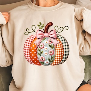 Könnte beinhalten: Cremefarbenes Sweatshirt mit einem bunten Kürbisdesign. Der Kürbis ist mit rot-weißem Vichy-Karo, orangefarbenen Punkten und grün-orangen Vichy-Mustern segmentiert. Eine rosa Schleife und Blumenstickerei zieren die Mitte, mit grünen Ranken und Blättern.