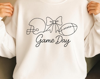 May include: White sweatshirt with a black graphic of a football helmet, bow, and football. The words "Game Day" are written in a cursive font below the graphic. The sweatshirt is a casual style.
