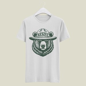 May include: White t-shirt with a black and white illustration of a bear wearing a ranger hat with the word "RESIST" on it.