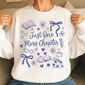 以下が含まれることがあります： 「Just One More Chapter」のテキストと、本、ハート、星、花、メガネ、リボンのイラストが描かれた、青いグラフィックデザインの白いスウェットシャツ。読書好きのための快適な衣服です。
