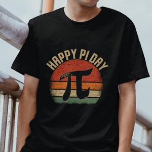 May include: Black t-shirt with a vintage-style graphic of a sunset with the text "Happy Pi Day" and a large pi symbol.
