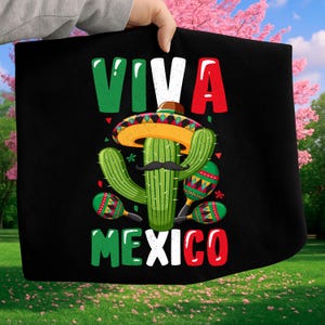 Puede incluir: Tela negra con un gráfico colorido que presenta un cactus con sombrero y bigote, con maracas a cada lado. Las palabras "VIVA MEXICO" están impresas en verde, blanco y rojo.