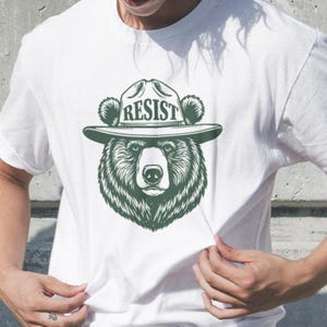 May include: White t-shirt with a green illustration of a bear wearing a ranger hat with the word "RESIST" on it.