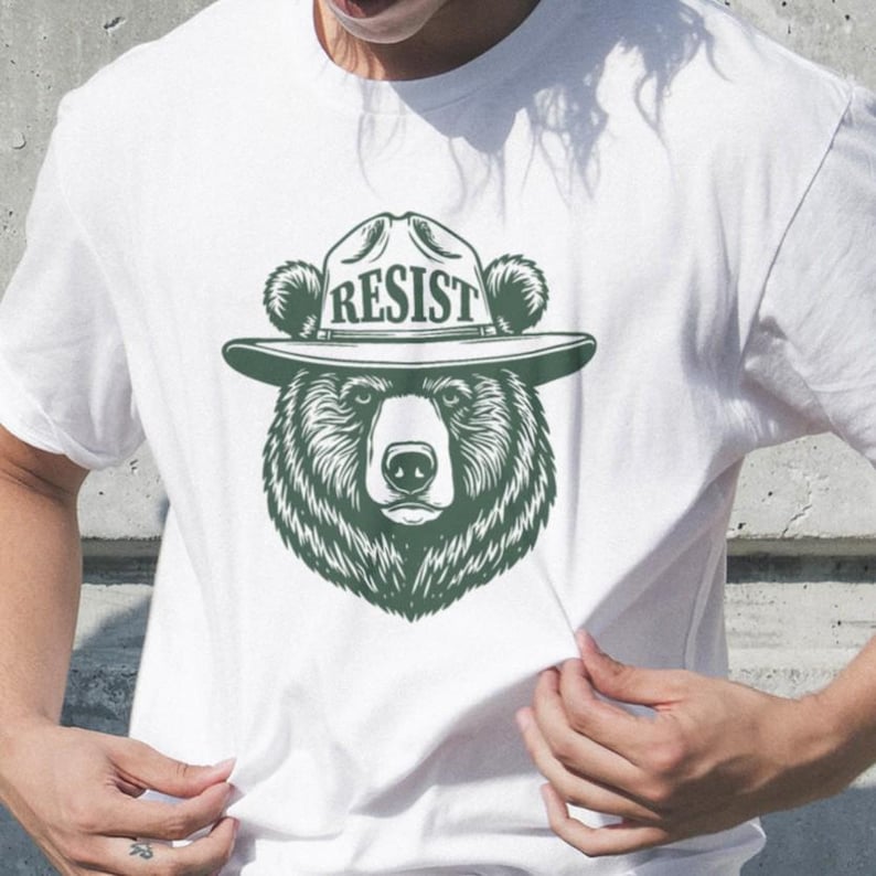 May include: White t-shirt with a green illustration of a bear wearing a ranger hat with the word "RESIST" on it.