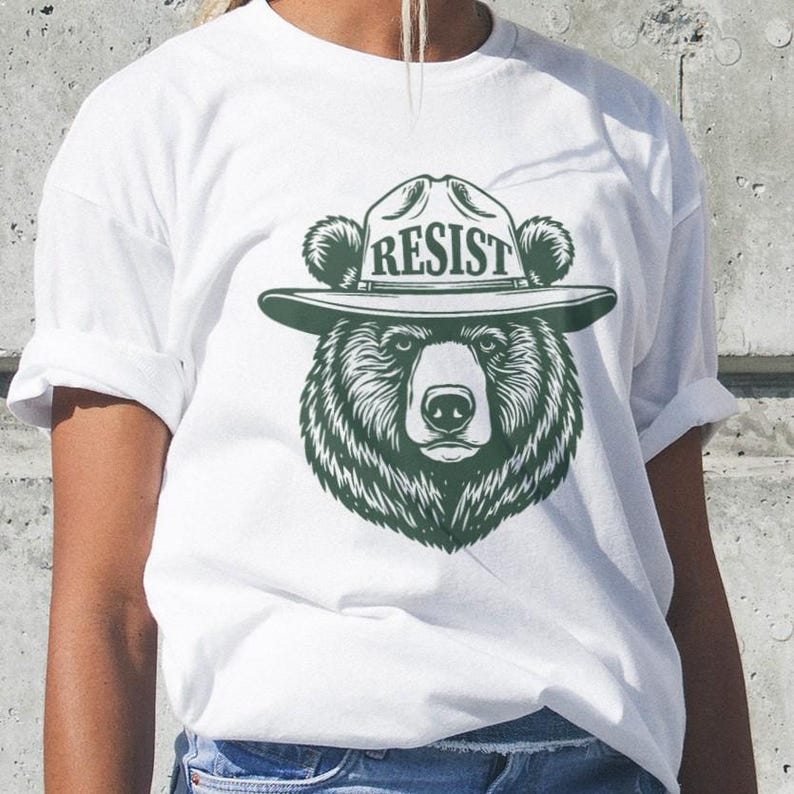 May include: White t-shirt with a green illustration of a bear wearing a ranger hat with the word "RESIST" on it.