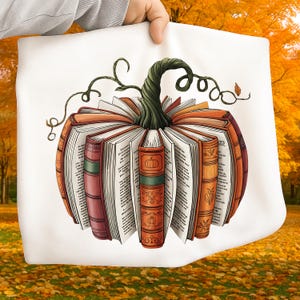 May include: A white tote bag featuring a pumpkin design made of open books. The books are orange and brown, with a green stem and vines. The background is a blurred autumn scene with yellow and orange leaves.