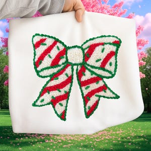 May include: A white fabric item featuring a large, embroidered bow design. The bow has a cream-colored center and is outlined in green. Red stripes and green speckles adorn the bow's loops, creating a festive, holiday-themed aesthetic.