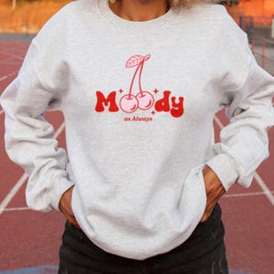 May include: A gray sweatshirt with a red and pink cherry graphic and the text "Moody as Always".