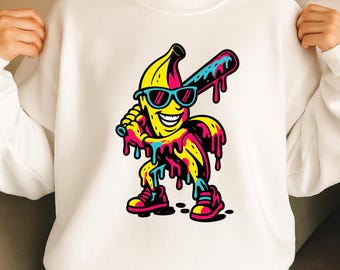 May include: White sweatshirt with a cartoon banana character wearing sunglasses and holding a baseball bat. The banana has a smiling face and is dripping with colourful paint. The design includes pink, yellow, blue, and black accents.