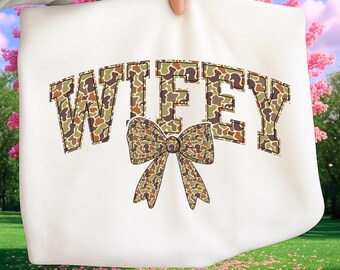 May include: White sweatshirt featuring the word "WIFEY" in a camouflage pattern, arched across the top. A camouflage bow is centered below the word. The sweatshirt is held up against a background of pink flowers and green grass.