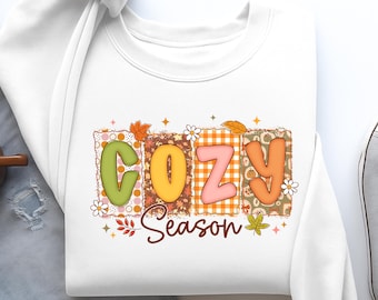 May include: White sweatshirt with the word "COZY" in colorful, bubble letters. The letters are set against a patchwork background with fall-themed patterns and the word "Season" below. The design includes leaves and floral accents.