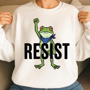 Puede incluir: Sudadera blanca con una rana verde con una bandana azul, levantando el puño. La palabra "RESIST" está impresa en letras negras en negrita debajo de la rana. La sudadera es de estilo casual y cómodo.