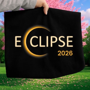 May include: Black tote bag with the word "ECLIPSE" in a bold font, and the year "2026" below. The word "ECLIPSE" is partially encircled by a glowing orange crescent. The bag is held up against a backdrop of a green lawn and pink flowering trees.