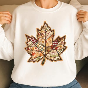 Könnte beinhalten: Weißes Sweatshirt mit einem großen, detaillierten Ahornblatt-Design. Das Blatt besteht aus verschiedenen Patchwork-Mustern in Herbstfarben wie Orange, Rot, Grün und Braun, was eine gemütliche, saisonale Ästhetik erzeugt.