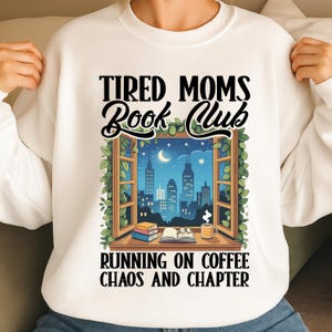Op de afbeelding: Witte sweatshirt met de tekst "Tired Moms Book Club" boven een afbeelding van een raam met uitzicht op een nachtelijke skyline. De tekst "Running on Coffee Chaos and Chapter" staat eronder.