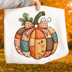May include: A white tote bag featuring a colorful patchwork pumpkin design. The pumpkin has sections of orange, brown, and teal fabric with floral and polka dot patterns. A plaid bow and green stem complete the fall-themed design.