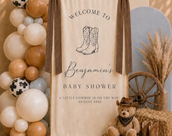 May include: A cream-colored fabric banner hanging from a wooden rod with brown ribbon bows. The banner features the text "WELCOME TO", "Benjamin's", "BABY SHOWER", "A LITTLE COWBOY IS ON THE WAY!", "AUGUST 2026", and a black line drawing of two cowboy boots.