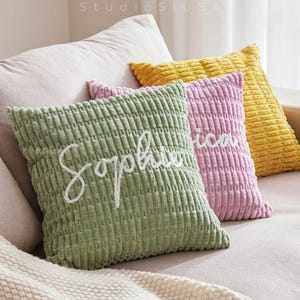 Puede incluir: Tres cojines de pana verdes, rosas y amarillos descansan sobre un sofá beige. El cojín verde tiene el nombre "Sophie" bordado en blanco. Los cojines añaden un toque de color al espacio de vida de tonos neutros.