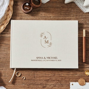 May include: A cream-colored wedding guest book with the initials "A" and "M" inside a brown floral design. The names "Anna & Michael" and the wedding date are printed below. The book is on a wooden surface with wedding rings and a pen.