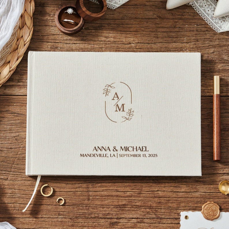 May include: A cream-colored wedding guest book with the initials "A" and "M" inside a brown floral design. The names "Anna & Michael" and the wedding date are printed below. The book is on a wooden surface with wedding rings and a pen.