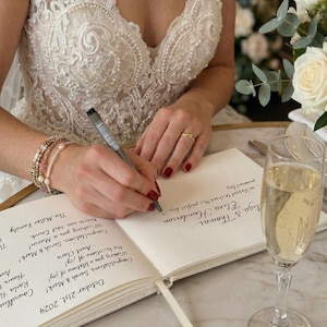 May include: A bride writing in a guest book at a wedding reception. The book is open, with handwritten messages and the date, October 21st, 2024. A glass of champagne sits nearby. The bride wears a white lace dress and jewelry.