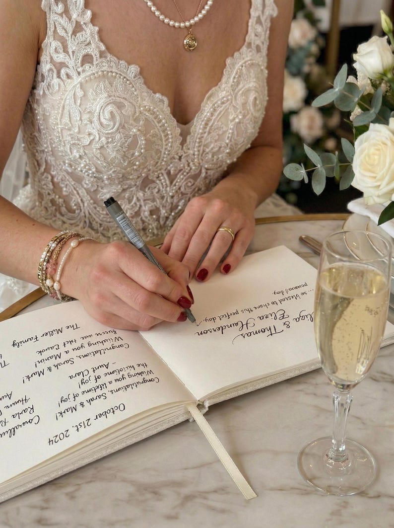 May include: A bride writing in a guest book at a wedding reception. The book is open, with handwritten messages and the date, October 21st, 2024. A glass of champagne sits nearby. The bride wears a white lace dress and jewelry.