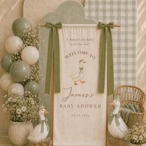 May include: A vertical fabric banner hanging from a wooden stand, featuring the text 'A Sweet Little Baby is on the way! WELCOME TO James's BABY SHOWER 08.09.2026' and a duck illustration. Two green fabric bows adorn the top corners of the banner.