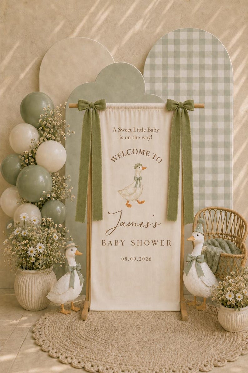 May include: A vertical fabric banner hanging from a wooden stand, featuring the text 'A Sweet Little Baby is on the way! WELCOME TO James's BABY SHOWER 08.09.2026' and a duck illustration. Two green fabric bows adorn the top corners of the banner.