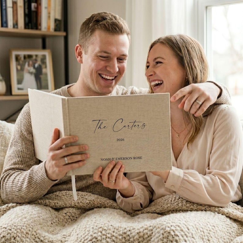 May include: A beige linen photo album with the text "The Carters 2026" and the names "Noah & Emerson Rose" is held open by a smiling couple. The album is on a patterned blanket, with a framed photo and a bookshelf in the background.