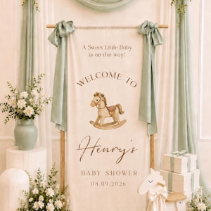 May include: A vertical fabric banner featuring the text A Sweet Little Baby is on the way! WELCOME TO Henry's BABY SHOWER 08.09.2026 with a watercolor rocking horse illustration, suspended from a wooden rod with sage green fabric drapes.