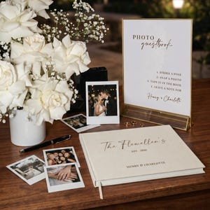 May include: A wedding guestbook and photo display featuring a sign with instructions, a book, and instant photos. The sign reads "PHOTO guestbook" and includes the names "Henry & Charlotte". A vase of white roses and a pen are also present.