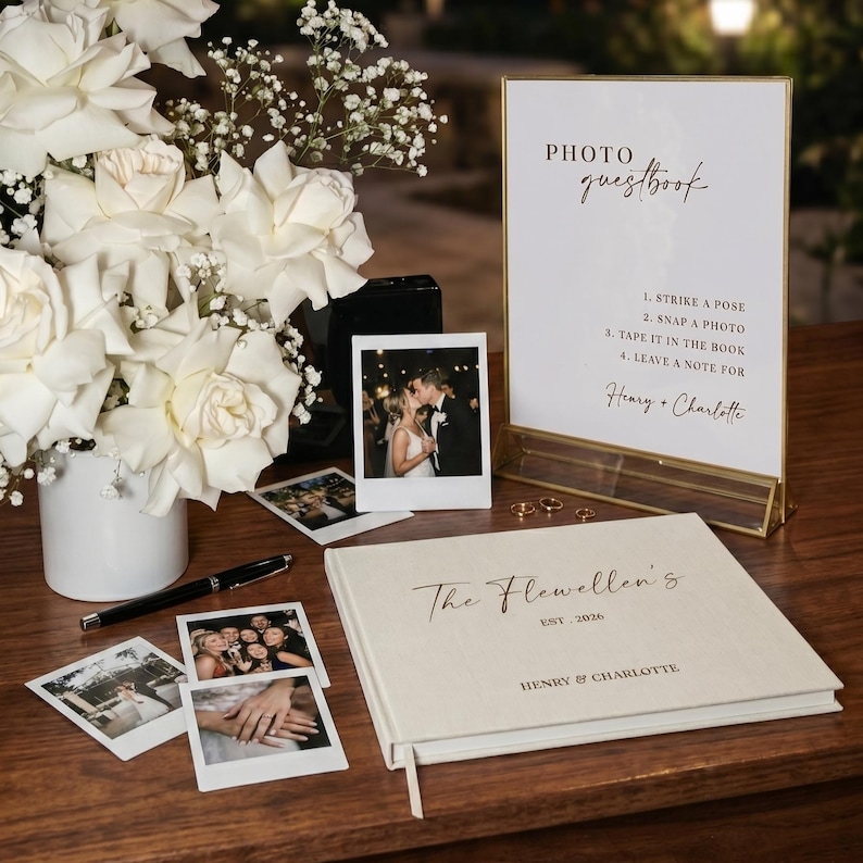 May include: A wedding guestbook and photo display featuring a sign with instructions, a book, and instant photos. The sign reads "PHOTO guestbook" and includes the names "Henry & Charlotte". A vase of white roses and a pen are also present.