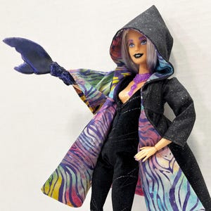 May include: A doll in a black jumpsuit and a hooded black coat with a colorful patterned lining. The doll has purple boots and a purple necklace. A large, dark blue, claw-like accessory extends from the doll's arm.