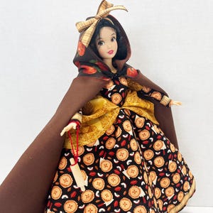 Op de afbeelding: Een pop met zwart haar draagt een bruine cape met capuchon en een geruite strik, een geel getextureerd accent in de taille en een zwarte jurk met een herhalend patroon van kleine appeltaartjes en rode appelschijfjes, met een miniatuur houten deegroller.