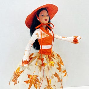 May include: A doll with long black hair wearing an orange wide-brimmed hat and a white long-sleeved top with orange leaf patterns. The outfit includes an orange bodice with a white band and a sheer skirt featuring metallic orange leaf motifs.