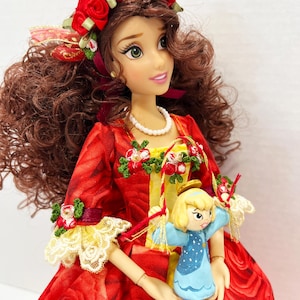 May include: A doll with brown curly hair, green eyes, and a red rose crown. The doll wears a red dress with a rose pattern, lace trim, and holds a small blue angel figurine. A pearl necklace adorns the doll's neck.