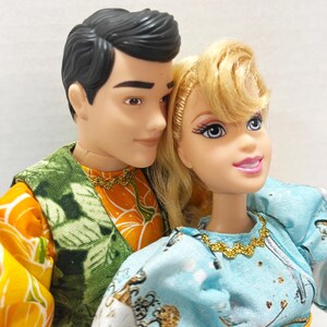 以下が含まれることがあります： 2体のファッションドール、男性と女性が寄り添っています。男性は黒髪で、緑とオレンジの模様のベストを着ています。女性はブロンドの髪で、金色のネックレスが付いた水色のドレスを着ています。